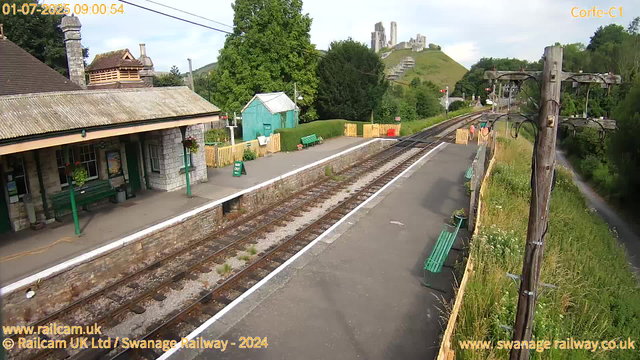 A small, rural train station with a platform on the left side of the image. There is a stone building with a tiled roof and hanging flower baskets. Near the building, there is a green bench and a "Way Out" sign. The train tracks run through the center of the image, curving slightly into the distance. On the right side, there are utility poles with wires, a narrow grass-covered area with wildflowers, and a pathway. In the background, a lush, green hillside features a castle ruin atop a hill, with some scaffolding and scaffolding supports visible. The sky is partly cloudy.