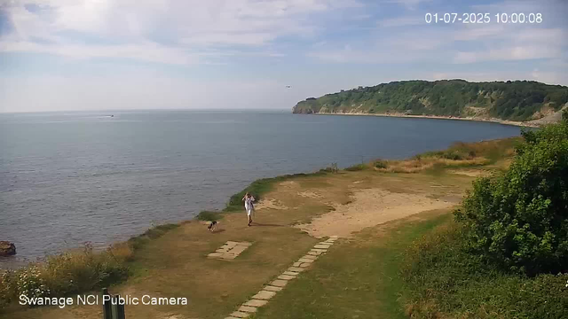 A person is walking along a grassy path by the water with a small dog. The scene includes a calm sea, distant cliffs covered in green vegetation, and a partly cloudy sky. The timestamp shows the date as July 1, 2025, at 10:00 AM.