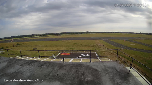 An outdoor view from a webcam at Leicestershire Aero Club shows a cloudy sky with some sunlight breaking through. In the foreground, there is a small parking area with a few white marked spaces and a red circle with a black 'X' on the asphalt, possibly indicating a no-parking zone. A metal railing borders the parking area. Beyond that, there is a grassy field with a wooden fence and a small roller or dolly. In the background, a runway extends across the scene, with the landscape beyond featuring trees and open fields. The image is timestamped "01-07-2025 10:00:11".