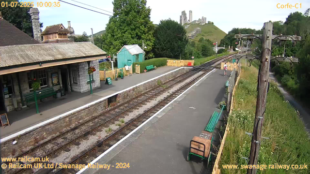 A small train station with a platform, a building with a sloped roof, green benches, and flower pots. A person with a backpack is waiting near the tracks. There are utility poles and wires along the side of the platform, with lush green trees and grass beyond. In the background, on a hill, there are ruins of an old castle. The sky is clear, and the scene is bright with daylight.