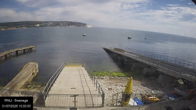 A view of a calm harbor with boats floating on the water. A concrete pier extends into the sea, with a sliding ramp or jetty at the end. There are railings along the walkway and some algae-covered rocks at the base of the pier. In the foreground, there are yellow caution signs and equipment, including a wheelbarrow and containers. The background features a coastline with cliffs and hills under a partly cloudy sky.