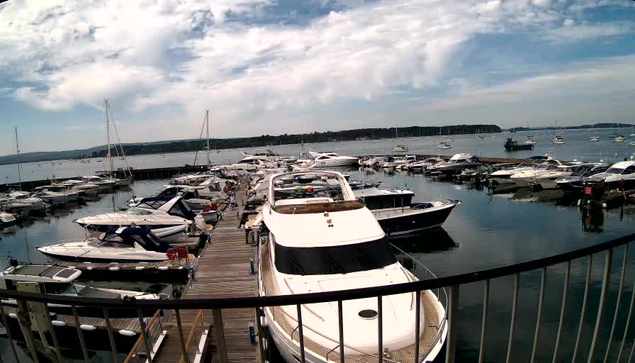 A marina filled with numerous docked boats and yachts on calm water, with a wooden pier leading into the distance. The sky is partly cloudy with patches of blue. A curved railing is visible in the foreground.
