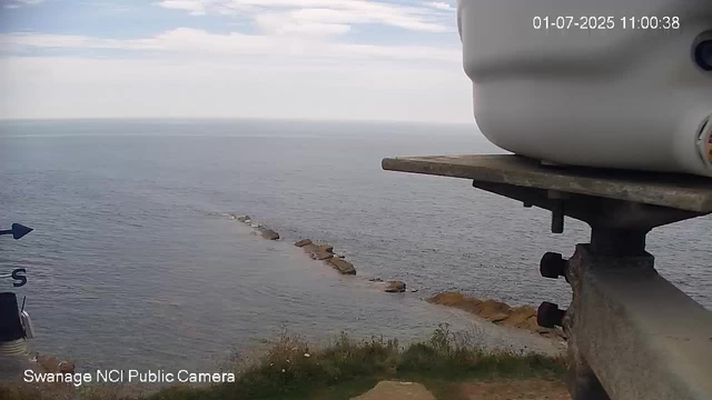 This is a view from a webcam showing a body of water with a rocky shoreline extending into the distance. The sky above is partly cloudy, and the water appears calm. There is some marine equipment, labeled "Swanage NCI Public Camera," visible in the lower left corner. In the upper right corner, the date and time are displayed as "01-07-2025 11:00:38." On the right side, part of a large white device and a wooden plank mounted on a metal support are visible in the foreground.
