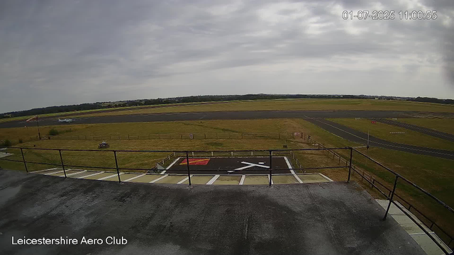 A view from a webcam at Leicesterhire Aero Club showing a cloudy sky over a runway and grassy field. In the foreground, there is a helicopter landing pad with a red and white marking. On the grassy area, there is a small vehicle and a flag on a pole. A small airplane is on the runway. The scene is framed by safety railings, and the timestamp in the top right corner reads "01-07-2025 11:00:55".