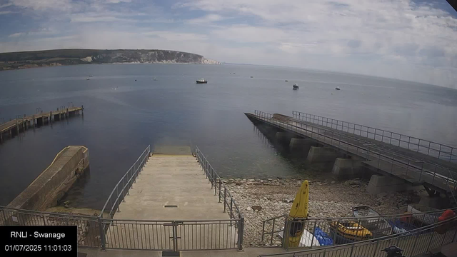 A view of a calm seaside scene with a pier extending into the water on the left and boats floating on the water. There is a concrete dock with railings in the foreground, along with a yellow buoy and some kayak or small boat equipment. In the background, a large hill or cliff outline is visible under a partly cloudy sky.