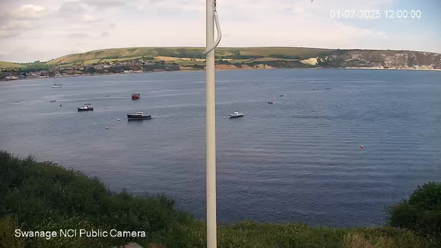 An image of a coastal scene viewed from a webcam. The water is calm with several boats floating, including small motorboats and a sailboat in the distance. There are buoys scattered across the water. In the background, there are green rolling hills and white chalk cliffs along the shoreline under a partly cloudy sky. A white pole is centered in the foreground. The date and time are displayed in the top right corner, showing January 7, 2025, at noon. The bottom left corner indicates that the image is from the Swannage NCI Public Camera.