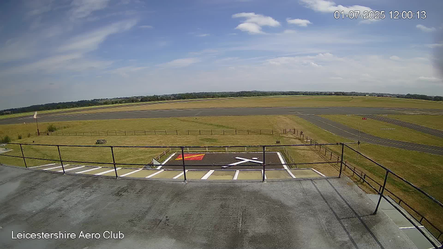 An outdoor scene at Leicesterhire Aero Club showing a clear sky with some scattered clouds. In the foreground, there is a parking area with clearly marked white lines and a red and yellow marked designated area, along with a railing and a walkway. Beyond that, a fenced grassy area extends to a runway, with some small runway markings visible. In the background, there are fields and trees stretching toward the horizon. The timestamp shows the date as July 1, 2025, at 12:00:13.