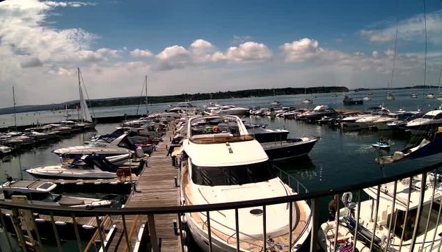 A marina filled with numerous boats and yachts docked along wooden piers. The water is calm, and there are clouds scattered across a bright blue sky. The photo appears to be taken from a balcony or viewing area with a black railing in the foreground.