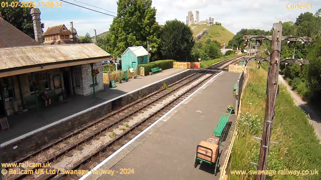 A rural train station platform on a sunny day, with an old stone building on the left side, benches, and flower baskets hanging on the building. The platform runs alongside railway tracks, and there is a yellow fence at the far end. To the right, tall wooden poles with electrical wires run parallel to the tracks, and there is greenery with grass and bushes growing next to the platform. In the background, a hill with ruins or castle remains is visible, and the sky is partly cloudy. A small turquoise shed and some small objects are also on the platform.