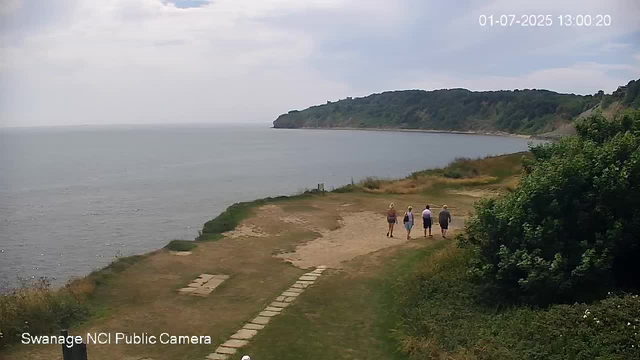 Four people are walking along a coastal path near a body of water, with a large wooded hill in the background. The sky is partly cloudy, and the scene appears calm and serene. The image is from a webcam, with the date and time displayed in the upper right corner.