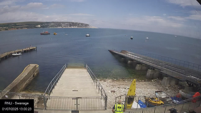 A seaside view shows a small harbor with a concrete dock extending into calm blue water, surrounded by a metal railing. To the right, there’s a larger ramp leading into the water with a safety railing. A person wearing a high-visibility vest stands in the foreground, facing the water. Nearby are yellow safety signs, a small boat, and some equipment. Boats are anchored in the water, with a large cliff in the background under a partly cloudy sky. The image is timestamped July 1, 2025, at 1:00 PM.