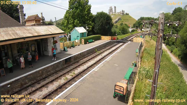 A small train station platform with a stone building on the left, where several people are waiting. There are benches, flower pots, and a green shed at the end of the platform. Railway tracks run through the center of the image, with a canopy and overhead electrical wires supported by wooden poles on the right. In the background, a lush, green hillside has an old castle ruin on top. The sky is partly cloudy.