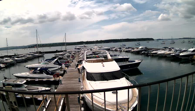 A view of a marina with multiple boats docked along a wooden pier. The boats vary in size, with some large yachts and smaller motorboats. The water is calm and reflects the boats and sky. In the background, there is a cloudy sky and a distant shoreline with a few boats sailing far out at sea. The perspective is from a vantage point behind a black metal railing.