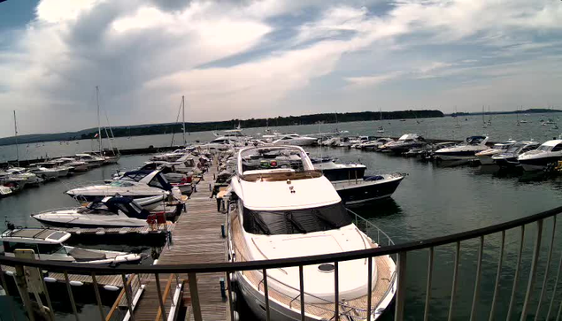 A harbor filled with numerous docked boats and yachts, with a wooden pier extending into the water. The sky is partly cloudy with hints of sunlight breaking through, and a distant shoreline is visible on the horizon. A safety railing curves along the foreground of the scene.