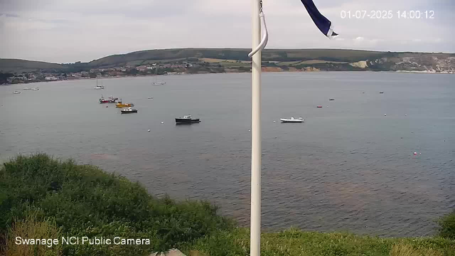 A view of a harbor with several boats anchored on calm water. In the foreground, there are green bushes, and a white flagpole with a flag. The background shows rolling hills and a cloudy sky. The timestamp in the upper right corner indicates the date and time as July 1, 2025, at 14:00:12.