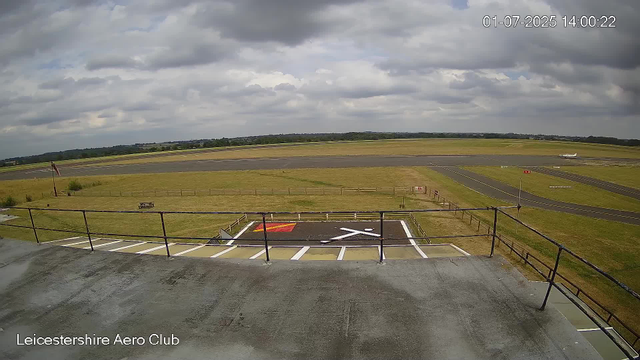 A view of an airfield shows a parking area in the foreground with a marked helicopter landing pad. Beyond the parking area, there are grassy fields, some pathways, and a runway. A small airplane is parked on the runway towards the right side of the image. The sky is mostly cloudy with patches of sunlight breaking through. The image is timestamped "01-07-2025 14:00:22" and has the label "Leicestershire Aero Club" at the bottom left corner.