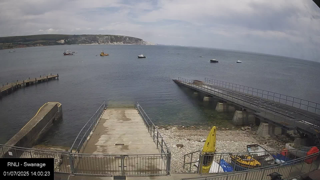 A view of a harbor with a concrete pier extending into calm water, bordered by metal railings. Several small boats are anchored in the water, with cliffs and a cloudy sky in the background. On the right side of the image, there is a raised pathway or breakwater also with railings, supported by concrete bases. In the foreground, there are some equipment and safety items, including a yellow life buoy and a yellow kayak, along with other miscellaneous objects near a fenced area. The timestamp indicates the date and time as July 1, 2025, at 14:00.