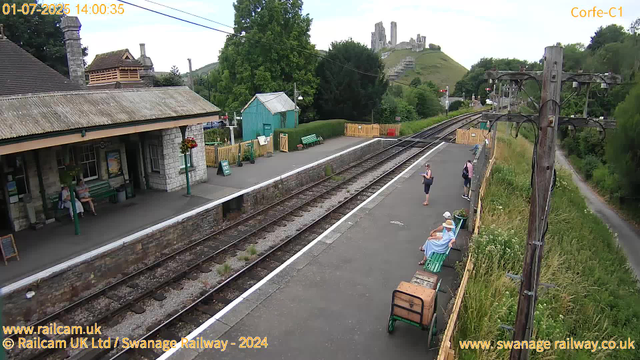 A small railway station platform with a few people waiting. Two women are seated on benches, one wearing a sunhat and a light-colored dress, the other in casual clothing. Three other women are standing near the edge, one reading a phone. The station building, made of stone, is on the left, with hanging flower baskets and a green bench outside. A narrow rural path runs alongside the right side with a grassy embankment behind. In the background, a lush green hillside features the ruins of a castle, and there are electrical poles and wires overhead. The weather appears clear and sunny.
