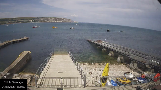 A view of a harbor with several boats floating on the water. In the foreground, there is a small dock with railings, a pebbled area, and some equipment including a yellow kayak and life jackets. The background features cliffs and a partly cloudy sky.
