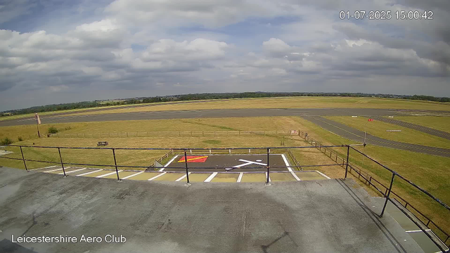 A wide view of an airport runway and surrounding grassy areas, taken from a webcam attached to a building at Leicestershire Aero Club. The sky is partly cloudy with patches of blue, and the scene appears calm and empty with no visible aircraft or people. The timestamp in the upper right corner shows the date as July 1, 2025, and the time as 15:00:42.