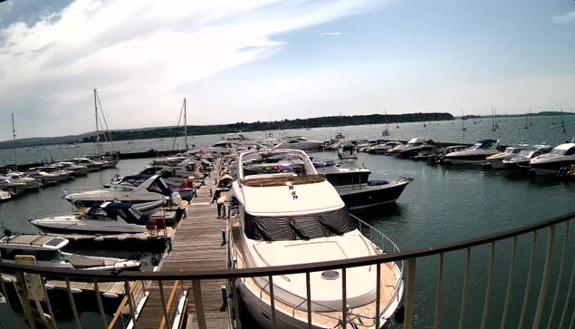 A marina filled with various boats and yachts docked along piers extending into calm water. The scene is set on a partly cloudy day with a low horizon in the background. A curved metal railing is visible in the foreground.