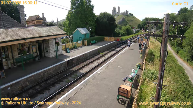 A small train station platform with a few people waiting, some sitting on benches and others standing near the edge. There is a woman wearing a hat sitting on a bench, with an old-fashioned luggage cart beside her. The station building has a stone facade and a roof, with hanging flower baskets and a green bench outside. A small green shed is also visible. The railway tracks run parallel to the platform, and on the right side, tall wooden electric poles with wires stretch across the scene. In the background, there is a lush green hillside with a castle ruin atop it. The weather is sunny, and trees and bushes line the surroundings.