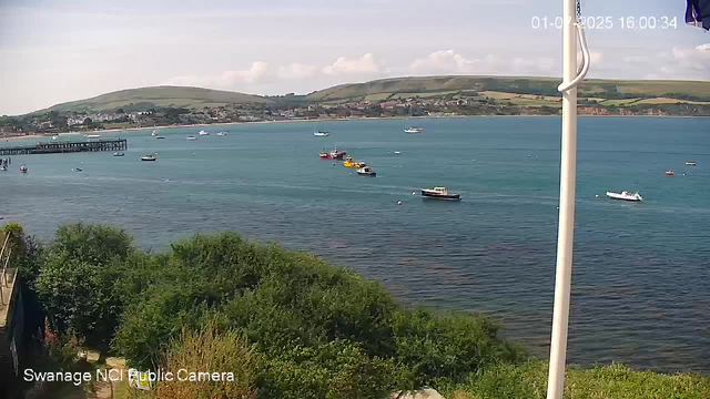 A view of a calm harbor with several boats floating on the water, some moored and some sailing, with green hills and scattered houses in the background. There is a pier on the left side and some trees in the foreground. A partial flagpole with a flag is visible on the right side of the image. The timestamp in the top right corner shows the date and time.