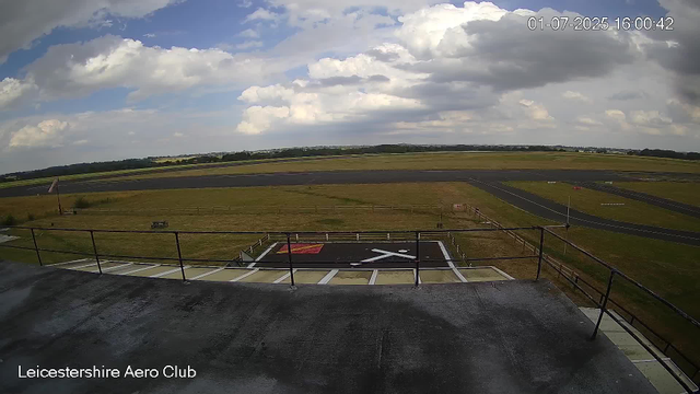 A view from a webcam at Leicestershire Aero Club showing a mostly cloudy sky with some blue patches, a wide grassy airfield with taxiways, a helicopter landing pad with a white "X" mark, and a small fencing area in the foreground. There are no people or aircraft visible, and the scene reflects a daytime setting.