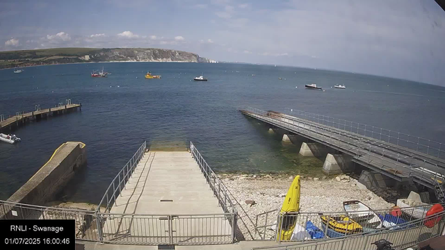 A view of a harbor with several boats floating on the water under a partly cloudy sky. In the foreground, there is a concrete dock area with a metal railing, a small boat, and some yellow and orange safety equipment, including a yellow kayak and life vests. To the right, a ramp extends into the water, supported by concrete supports. In the background, there are distant cliffs and green land across the water. The timestamp indicates the date and time as July 1, 2025, at 4:00 PM.