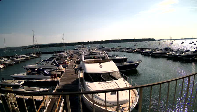 A marina with numerous boats docked along wooden piers. The harbor is filled with various sized yachts and smaller boats, with a calm water surface sparkling in the sunlight. A curved railing is visible in the foreground, and a distant shoreline stretches across the background under a partly cloudy sky.