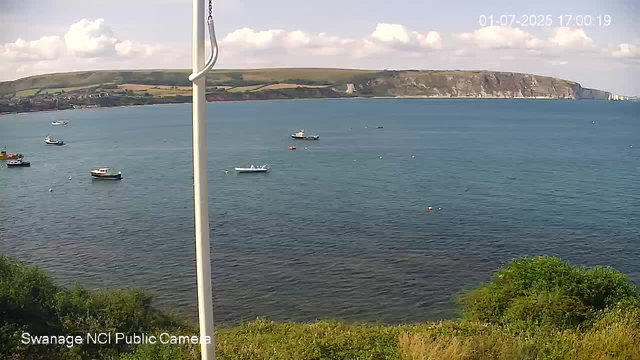 A view of a coastal area featuring calm blue water with several small boats floating on the surface. In the background, there are green hills with partly cloudy skies overhead. A white pole partially obstructs the view in the foreground. The image has a timestamp showing July 1, 2025, at 17:00:19. The location is identified as Swannage NCI Public Camera.