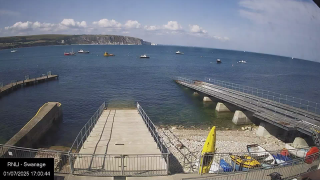 A view of a harbor with several boats floating on the water. There are two piers extending into the sea, one on the left and one on the right. In the lower part of the image, there is a small area with a yellow lifeguard rescue board, some parked vehicles, and fencing. The sky is mostly clear with some scattered clouds, and a cliff is visible in the distance across the water.