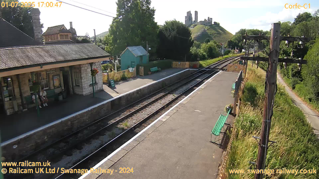 A small train station platform with a stone building on the left, where a person is sitting on a bench. There are flower baskets hanging near the entrance. A sign indicates the direction for exiting the station. Railway tracks run through the center, with some signal equipment visible. On the right, there are green benches along a fence, with tall grass and bushes beyond it. In the background, a hill with a castle ruin at the top is visible, along with trees and a clear sky. The timestamp indicates the photo was taken on July 1, 2025, at 5:00 pm.