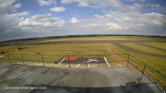 A view from a webcam at Leicestershire Aero Club showing a clear sky with scattered clouds. The foreground features an empty helipad with a red and black marking, surrounded by a railing, and a bench nearby. Beyond the helipad, there are grassy areas, a taxiway, and open fields extending to the horizon. The top right corner displays the date and time as 01-07-2025 17:00:45.