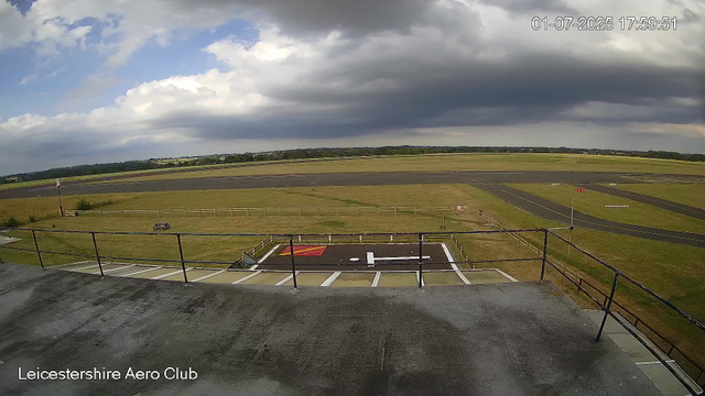 A wide view of an airfield with runways and grassy areas under a partly cloudy sky. The image is taken from a platform or balcony with a black railing in the foreground. The timestamp shows "01-07-2025 17:59:51" and the location is labeled "Leicestershire Aero Club" at the bottom left corner.