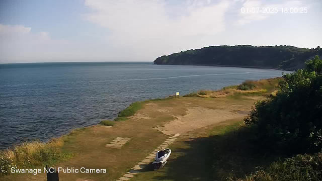 A scenic view of a coastal area with calm blue water stretching toward a distant tree-covered hill or headland. In the foreground, there is a small grassy or sandy area with a weathered bench, and a person lying on the bench facing upward, possibly resting or relaxing. The scene is bathed in sunlight, with some clouds in the sky, and the timestamp indicates the image was captured on July 1, 2025, at 6:00 PM. The bottom left corner shows the text "Swanage NCI Public Camera."