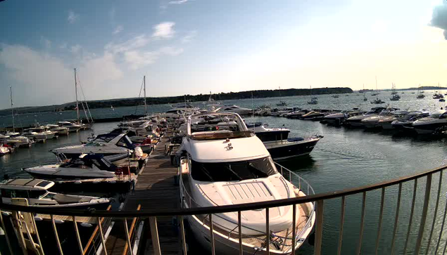 A marina filled with numerous yachts and boats docked along wooden piers, with calm water and a distant shoreline under a partly cloudy sky.