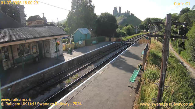 A small train station platform with a building on the left, featuring stone walls and a corrugated metal roof. There are benches and flower baskets hanging, and the platform is bordered by a low fence. To the right, there are overhead electric lines supported by wooden poles. The railway tracks run through the center, curving slightly into the distance. In the background, a lush green hillside with a castle or ruins atop it is visible under a clear sky, with the sunlight casting shadows on the platform.