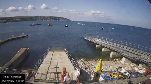 A view of the seaside at Swanage on a sunny day. The image shows a small pier extending into calm blue water, with several boats moored in the distance. There is a rocky shoreline and a gently sloping green hill or cliff in the background. In the foreground, three people are gathered near the railing, engaging in conversation, with one person holding a hat. A yellow rescue boat and some parked vehicles are also visible near the bottom right corner. The sky is mostly clear with a few fluffy clouds.