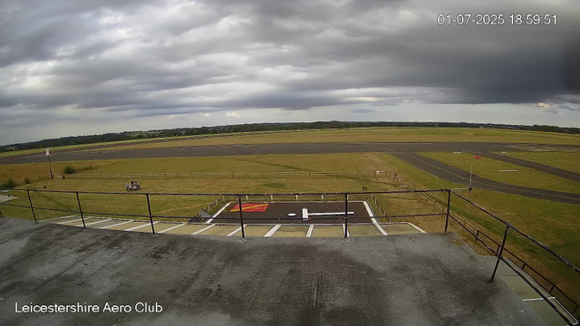 An outdoor view from a webcam at LeicesterShire Aero Club shows an expansive sky filled with dark, heavy clouds. Below the sky, there is a large grassy area with a small paved helipad marked with a white helicopter symbol. The helipad is enclosed by a low fence. In the distance, there are winding taxiways and runways, with some signage visible. A small, grassy open space includes two parked golf carts, and a windsock is positioned near the fence on the left side of the image. The scene appears to be at an airport or airfield, under overcast weather conditions.