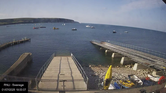 A view of a harbor with small boats anchored in the water, a pier extending into the sea, and a yellow lifeguard float near the shore. In the background, there is a distant shoreline with cliffs and green hills. The sky is mostly clear with a few clouds. The timestamp indicates the date as July 1, 2025, at 7:00 PM.
