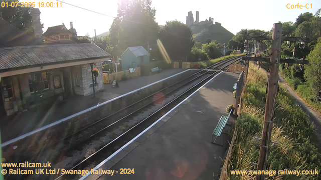 A small railway station platform with a single building on the left, and overgrown grass and bushes on the right. The train tracks run through the center of the image, curving slightly to the right in the distance. There are two green benches on the platform, one near the building and another further to the right. A wooden utility pole with wires stands on the right side of the platform. The background features a hillside with ruins or old structures at the top, surrounded by trees. The sun is low in the sky, casting bright light and creating lens flares. The image has date and time displayed at the top left corner, and website credits at the bottom.