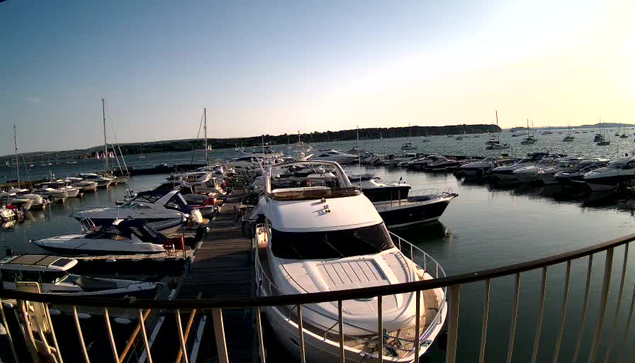 A marina filled with numerous boats and yachts docked along wooden piers. The water is calm and reflects the boats. The sky is clear with a soft light, suggesting it is either early morning or late afternoon. A curved railing is visible in the foreground, framing the scene.