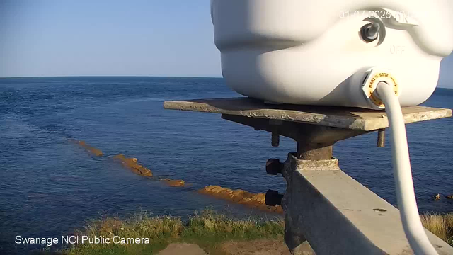 A view of the ocean with a rocky shoreline extending into the water. In the foreground, there is a mounted weather or camera device attached to a wooden and metal structure, with a white cable connected to it. The sky is clear and blue.