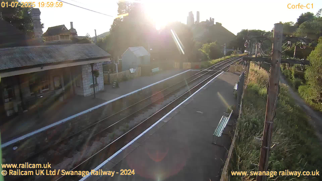 A small railway platform is shown bathed in the warm glow of the setting sun. There is a single bench on the platform, and a building with a stone facade and a corrugated metal roof on the left side. Overhead, there are electrical wires and poles running parallel to the train tracks, which are curving into the distance. In the background, a hill with a castle or fortress ruins is visible, surrounded by trees and greenery. There are also various small structures and fencing along the platform, with some benches and signs. The scene is peaceful, with the sunlight creating lens flare effects and casting long shadows across the platform.