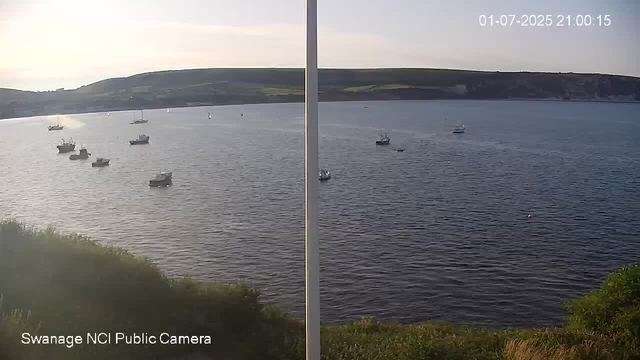 A split-screen image from a webcam shows a body of water with several boats anchored or sailing. The left side appears brighter with sunlight, while the right side is darker, indicating different lighting conditions or times of day. In the background, there are rolling hills with patches of greenery. The timestamp in the upper right corner reads "01-07-2025 21:00:15." At the bottom left, there is text that says "Swanage NCI Public Camera."