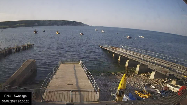 A view of a harbor with calm water, several small boats floating in the distance, a wooden dock extending into the water on the left, a concrete pier on the right, and a rocky shoreline at the bottom. There is a yellow caution sign and some vehicles parked near the shore, with a grassy hill and cliffs visible in the background under a clear sky.