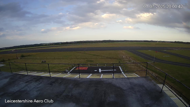 A view from a webcam at Leicestershire Aero Club showing an outdoor airfield scene with a runway stretching into the distance under a partly cloudy sky. The foreground features a small helipad marked with a white 'H' on a black surface, surrounded by a fenced area. A small bench is visible on the grass to the left, and flight information boards are positioned along the fence. The timestamp in the upper right corner indicates the date and time as January 7, 2025, at 8:00 PM.