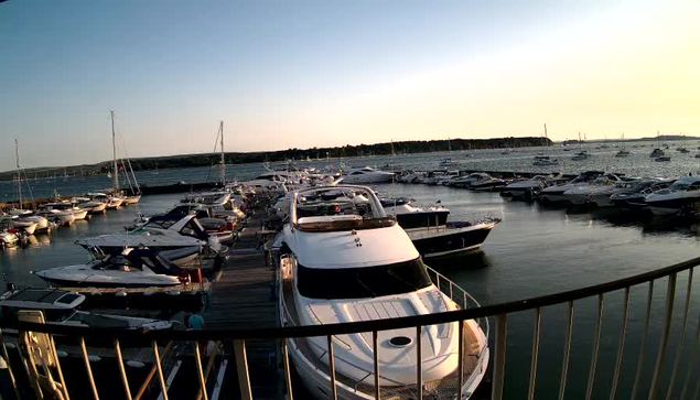 A marina filled with numerous boats and yachts docked along wooden piers, with calm water reflecting the vessels. The scene is set during sunset, casting a warm glow over the boats and the horizon. A curved metal railing is visible in the foreground.