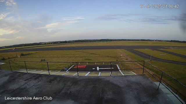 An empty outdoor area of Leicestershire Aero Club seen from a high vantage point. The foreground shows a rooftop with a railing, a helipad marked with a red square and a white "H," and some equipment. In the background, there are grassy fields, taxiways, and runways stretching towards the horizon under a partly cloudy sky during late evening or dusk. The timestamp in the upper right corner reads "01-07-2025 21:00:42."